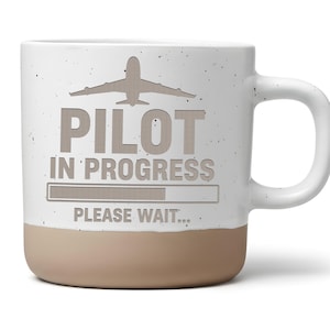 Könnte beinhalten: Ein weißer Keramikbecher mit gesprenkelter Oberfläche und beigem Boden. Die Vorderseite zeigt eine graue Flugzeuggrafik über dem Text PILOT IN PROGRESS, einem Ladebalken und dem Text PLEASE WAIT. Der Becher hat einen geschwungenen Henkel.