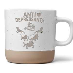 Könnte beinhalten: Ein weißer Keramikbecher mit gesprenkelter Oberfläche und einem beigen Boden. Auf der Vorderseite steht der Text ANTI DEPRESSANTS mit einem Herzsymbol, ergänzt durch Illustrationen von drei Pillendosen und vier Bartagamen in V-Form.
