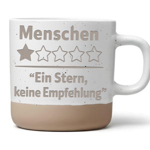 Könnte beinhalten: Ein weißer Keramikbecher mit gesprenkelter Oberfläche und einem beigefarbenen Boden. Auf der Vorderseite steht der Text "Menschen" über einer Fünf-Sterne-Bewertung mit einem ausgefüllten Stern, gefolgt von "Ein Stern, keine Empfehlung" in Grau.