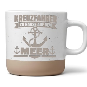 Könnte beinhalten: Ein weißer Keramikbecher mit Sprenkeloptik und beigem Boden. Auf der Vorderseite befindet sich eine graue Grafik eines Ankers mit Seil sowie der Schriftzug KREUZFAHRER ZU HAUSE AUF DEM MEER in grauen Blockbuchstaben über und unter dem Anker.