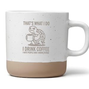 Könnte beinhalten: Weiße Keramik-Kaffeetasse mit beigefarbenem Boden und gesprenkelter Oberfläche. Die Tasse zeigt eine mürrische Schildkröten-Grafik, die eine Kaffeetasse hält, mit dem Text "THAT'S WHAT I DO, I DRINK COFFEE, I HATE PEOPLE AND I KNOW THINGS."