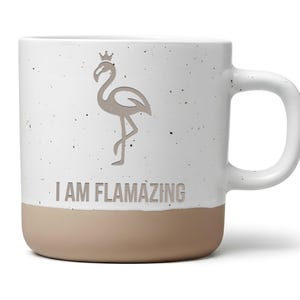 Könnte beinhalten: Ein weißer Keramikbecher mit gesprenkelter Oberfläche und einem beigefarbenen Boden. Der Becher zeigt eine graue Grafik eines Flamingos mit einer kleinen Krone sowie den Schriftzug I AM FLAMAZING in grauen Großbuchstaben auf der Vorderseite.