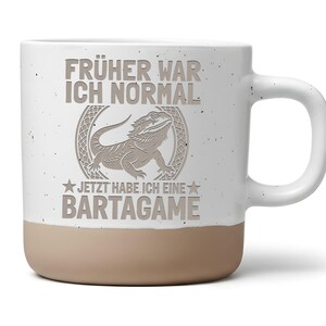 Könnte beinhalten: Ein weißer Keramikbecher mit gesprenkelter Oberfläche und beigem Boden. Auf der Vorderseite befindet sich eine Grafik einer Bartagame in einem kreisförmigen Rahmen sowie der graue Schriftzug "FRÜHER WAR ICH NORMAL JETZT HABE ICH EINE BARTAGAME".