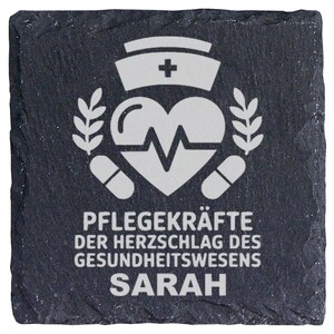 Könnte beinhalten: Ein quadratischer Untersetzer aus schwarzem Schiefer mit rauer, strukturierter Kante. In der Mitte befindet sich eine weiße Gravur mit einer Krankenschwesterhaube, einem Herz mit EKG-Linie, zwei Lorbeerzweigen und zwei Kapseln. Text: PFLEGEKRÄFTE DER HERZSCHLAG DES GESUNDHEITSWESENS SARAH.