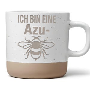 Könnte beinhalten: Ein weißer Keramikbecher mit gesprenkelter Oberfläche und einem beigefarbenen Boden. Auf der Vorderseite befindet sich eine braune Grafik einer Biene unter dem Text "ICH BIN EINE Azu-" in passendem Braunton. Der Becher hat einen seitlichen Henkel.