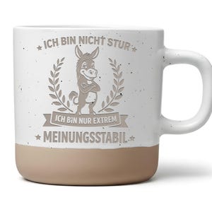 Könnte beinhalten: Ein weißer Keramikbecher mit Sprenkeloptik und beigem Boden. Auf der Vorderseite ist ein Esel mit verschränkten Armen abgebildet, umrahmt von einem Lorbeerkranz. Der Text lautet: ICH BIN NICHT STUR, ICH BIN NUR EXTREM MEINUNGSSTABIL.