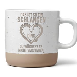 Könnte beinhalten: Eine weiße Keramiktasse mit Sprenkeloptik und beigem Boden. Auf der Vorderseite befindet sich eine graue Grafik einer Schlange in Herzform sowie der deutsche Text: DAS IST SO EIN SCHLANGEN DING DU WÜRDEST ES NICHT VERSTEHEN in grauer Farbe.