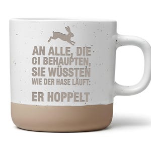 Könnte beinhalten: Ein weißer Keramikbecher mit Sprenkeloptik und beigem Boden. Der Becher zeigt eine Grafik eines springenden Hasen und den Text: AN ALLE, DIE BEHAUPTEN, SIE WÜSSTEN WIE DER HASE LÄUFT: ER HOPPELT. Der Becher verfügt über einen geschwungenen Henkel.