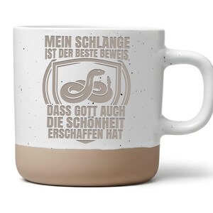 Könnte beinhalten: Ein weißer Keramikbecher mit Sprenkeloptik und beigefarbenem Boden. Auf der Vorderseite befindet sich eine graue Grafik einer Schlange in einem Schildemblem sowie der Text: MEIN SCHLANGE IST DER BESTE BEWEIS, DASS GOTT AUCH DIE SCHÖNHEIT ERSCHAFFEN HAT.