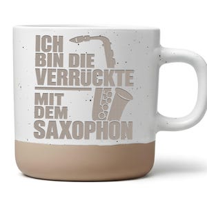 Könnte beinhalten: Ein weißer Keramikbecher mit Sprenkeloptik und beigefarbenem Boden. Auf der Vorderseite befindet sich eine braune Saxophon-Grafik und der deutsche Text "ICH BIN DIE VERRÜCKTE MIT DEM SAXOPHON" in einer fetten, serifenlosen Schriftart.