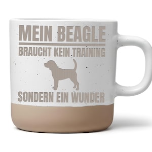 Könnte beinhalten: Ein weißer Keramikbecher mit gesprenkelter Oberfläche und einem beigefarbenen Boden. Auf der Vorderseite ist die Silhouette eines Beagle-Hundes sowie der Text MEIN BEAGLE BRAUCHT KEIN TRAINING SONDERN EIN WUNDER abgebildet. Der Becher hat einen Henkel.