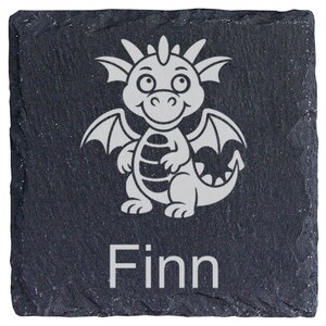 Könnte beinhalten: Ein quadratischer Untersetzer aus schwarzem Schiefer mit rauer, strukturierter Kante. In der Mitte befindet sich eine hellgraue Gravur eines Cartoon-Drachen mit Flügeln und Schwanz. Unter dem Drachen ist der Name Finn in einer serifenlosen Schrift eingraviert.