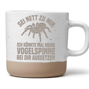 Könnte beinhalten: Ein weißer Keramikbecher mit Sprenkeloptik und beigefarbenem Boden. Auf der Vorderseite befindet sich eine Grafik einer Vogelspinne und der Text: SEI NETT ZU MIR. ICH KÖNNTE MAL MEINE VOGELSPINNE BEI DIR AUSSETZEN. Der Becher hat einen Henkel.
