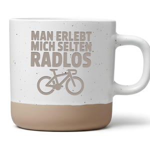 Könnte beinhalten: Weißer Keramikbecher mit gesprenkelter Oberfläche und beigefarbenem Boden. Der Becher zeigt den Text "MAN ERLEBT MICH SELTEN RADLOS" und eine Fahrradgrafik in Hellbraun. Der Becher hat einen großen, gebogenen Griff.