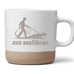 Könnte beinhalten: Weißer Keramikbecher mit beigefarbenem Boden und Henkel. Der Becher zeigt eine Grafik einer Person, die eine Eidechse an der Leine ausführt, mit dem Text ".exe ausführen" darunter.