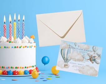 May include: A white birthday cake with colourful sprinkles and candles, beside a cream envelope and an invitation reading "Adventure Awaits!" with a hot air balloon and airplane illustration. A blue balloon and confetti are also present.