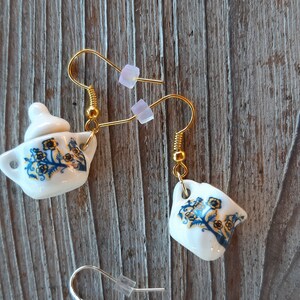 May include: White ceramic teapot earrings with gold-colored hooks. The earrings feature a blue and yellow floral design. One earring is a teapot, and the other is a teacup. The earrings are displayed on a wooden surface.