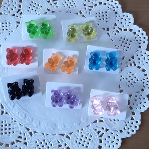 May include: A collection of colorful gummy bear earrings in various colors, including green, yellow, red, orange, blue, black, purple, and pink. Each pair is mounted on a small white card, displayed on a white lace doily.