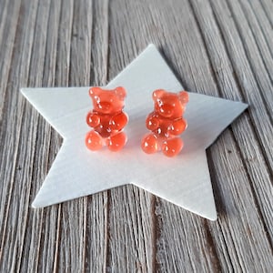 May include: Two translucent, red gummy bear candies sit atop a white star-shaped decoration. The bears are detailed with visible arms, legs, and ears. The background is a weathered, gray wooden surface.