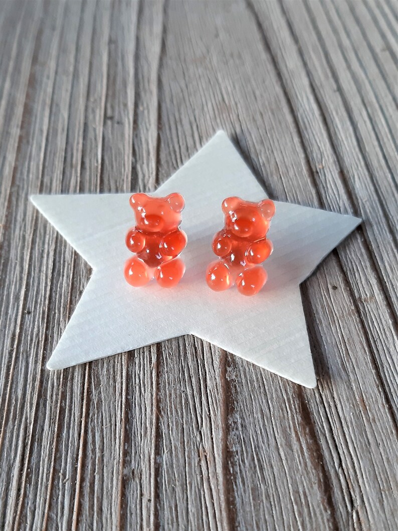 May include: Two translucent, red gummy bear candies sit atop a white star-shaped decoration. The bears are detailed with visible arms, legs, and ears. The background is a weathered, gray wooden surface.