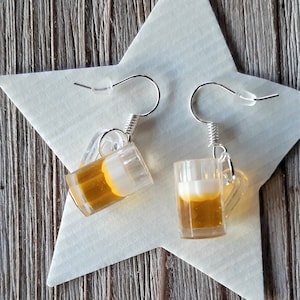 May include: A pair of novelty earrings featuring miniature beer mugs. The clear, rectangular mugs are filled with a golden-brown liquid and topped with a white foam. The earrings hang from silver-colored hooks and are displayed on a white star.