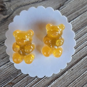 May include: Two gummy bear candies in a golden yellow color, positioned on a white, scalloped-edge paper doily. The candies are translucent, with visible details of their bear shape. The background is a wooden surface.