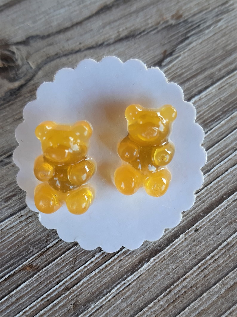 May include: Two gummy bear candies in a golden yellow color, positioned on a white, scalloped-edge paper doily. The candies are translucent, with visible details of their bear shape. The background is a wooden surface.