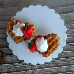 Puede incluir: Dos gofres en forma de coraz&oacute;n con crema batida y una fresa roja sobre un papel blanco festoneado. Los gofres son de color marr&oacute;n dorado y las fresas tienen tallos verdes. El fondo es una superficie de madera.