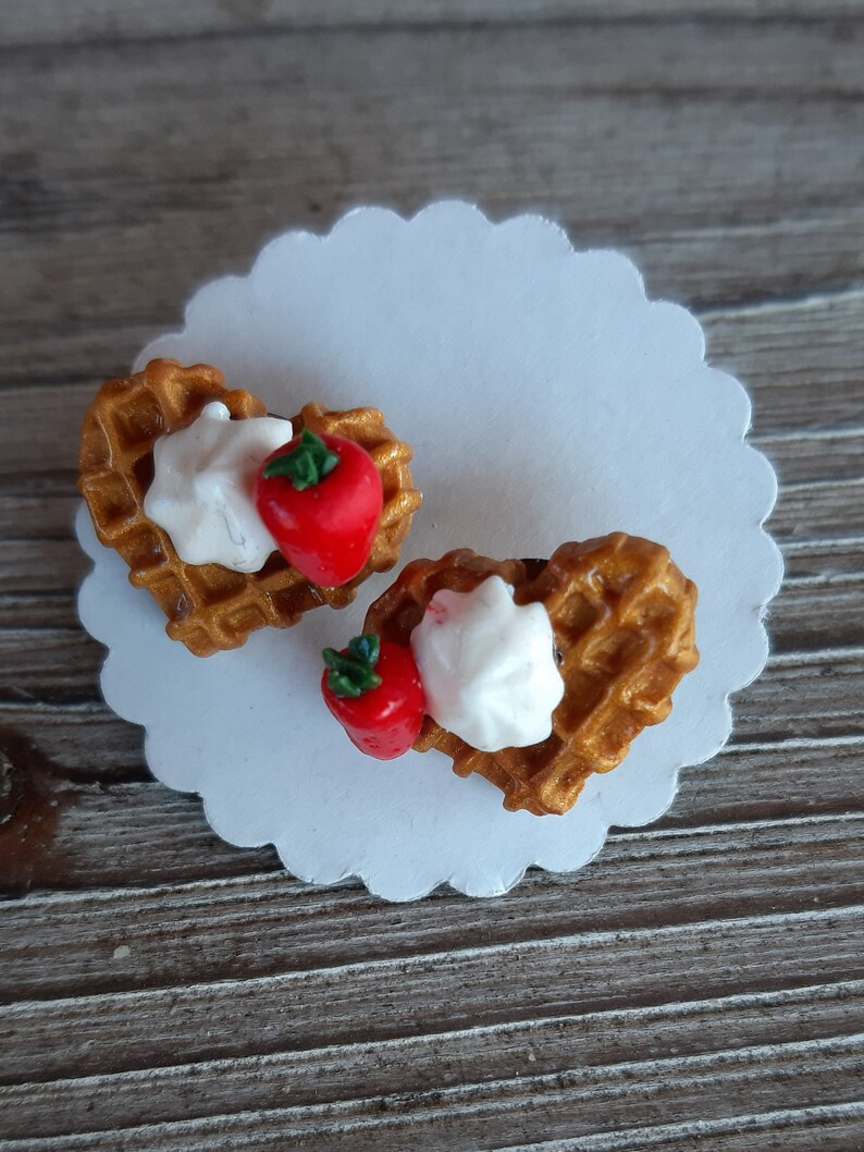 Puede incluir: Dos gofres en forma de coraz&oacute;n con crema batida y una fresa roja sobre un papel blanco festoneado. Los gofres son de color marr&oacute;n dorado y las fresas tienen tallos verdes. El fondo es una superficie de madera.