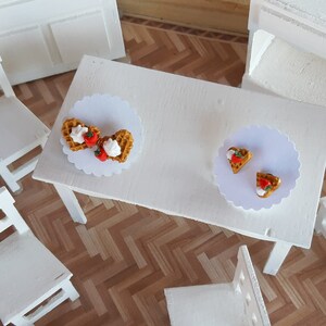 Puede incluir: Un juego de comedor blanco en miniatura con una mesa y sillas. La mesa est&aacute; decorada con dos blondas de papel blancas, cada una con un gofre cubierto con crema batida y una fresa. La escena est&aacute; ambientada en un suelo de parquet.