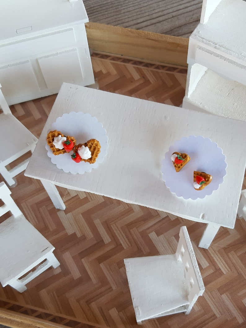 Puede incluir: Un juego de comedor blanco en miniatura con una mesa y sillas. La mesa est&aacute; decorada con dos blondas de papel blancas, cada una con un gofre cubierto con crema batida y una fresa. La escena est&aacute; ambientada en un suelo de parquet.