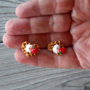 Puede incluir: Dos pendientes en miniatura con forma de gofre. Cada pendiente presenta una base de gofre, cubierta con crema blanca y una fresa roja. Los pendientes son peque&ntilde;os y detallados, con una apariencia realista. El fondo es una superficie de madera gris claro.