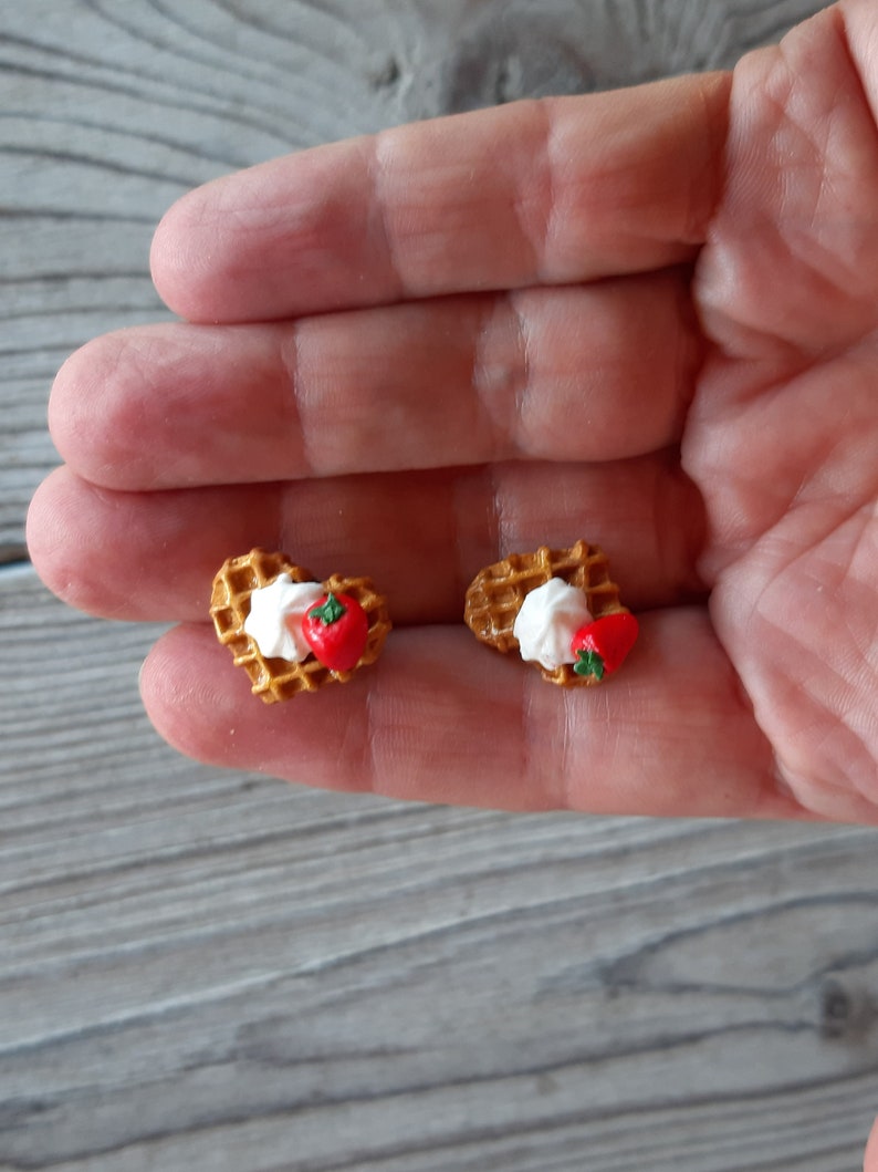 Puede incluir: Dos pendientes en miniatura con forma de gofre. Cada pendiente presenta una base de gofre, cubierta con crema blanca y una fresa roja. Los pendientes son peque&ntilde;os y detallados, con una apariencia realista. El fondo es una superficie de madera gris claro.