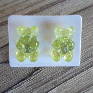May include: Two translucent, lime green gummy bear candies sit in a white plastic mold. The gummy bears are detailed with rounded features and are positioned on a wooden surface.
