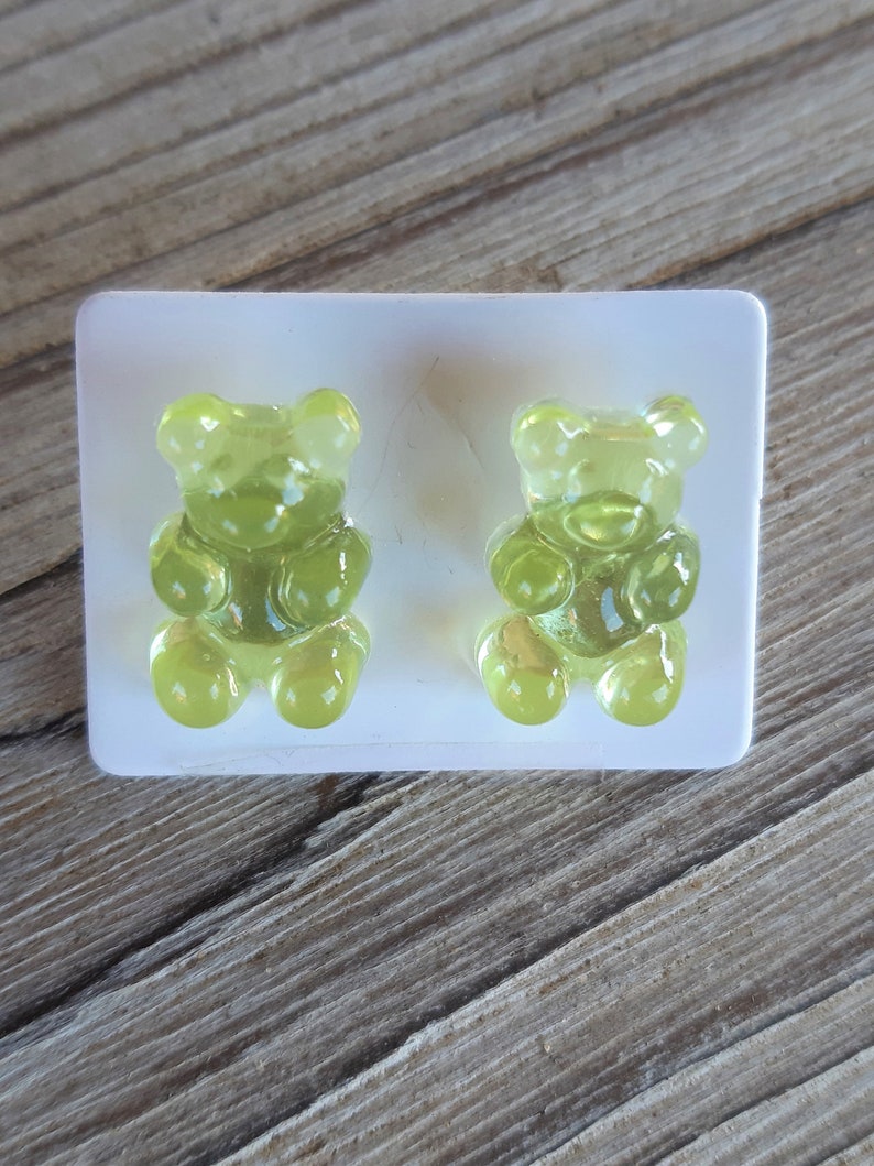 May include: Two translucent, lime green gummy bear candies sit in a white plastic mold. The gummy bears are detailed with rounded features and are positioned on a wooden surface.