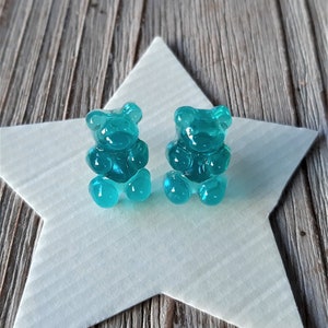 May include: Two teal gummy bear-shaped earrings sit on a white star-shaped paper cutout, against a weathered wood background. The earrings are translucent and have a glossy finish. The overall aesthetic is playful and whimsical.