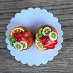Könnte beinhalten: Zwei Miniatur-Obsttörtchen auf einer weißen, mit Zacken verzierten Papierunterlage. Jedes Törtchen ist mit Kiwischeiben, Erdbeeren, Bananen und Orangenspalten sowie roten Beeren belegt. Die Törtchen liegen auf einer hellbraunen Holzoberfläche.