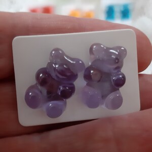 May include: A pair of purple gummy bear-shaped earrings. The earrings are a translucent lavender color and are mounted on a white rectangular card. Other colorful gummy bear earrings are visible in the background.