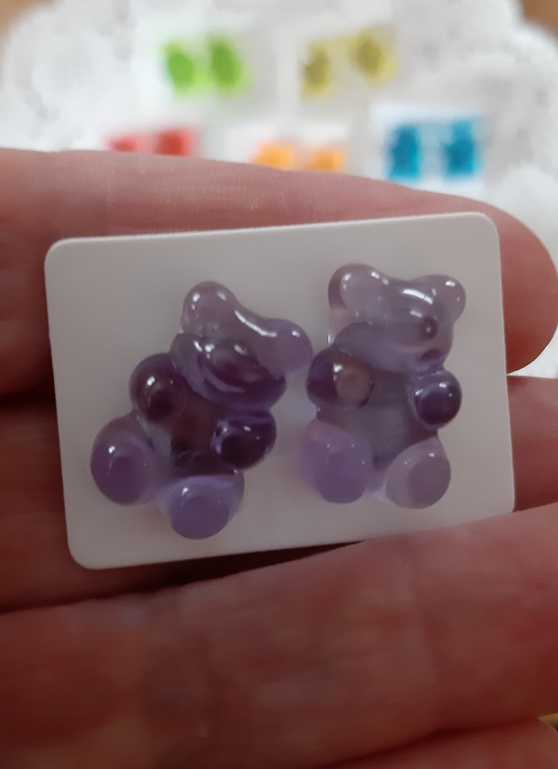 May include: A pair of purple gummy bear-shaped earrings. The earrings are a translucent lavender color and are mounted on a white rectangular card. Other colorful gummy bear earrings are visible in the background.