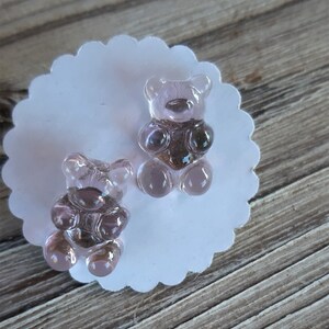 May include: Two translucent, light purple gummy bear-shaped charms sit on a white, scalloped-edge paper. The bears have a darker purple center and are positioned on a wooden surface.