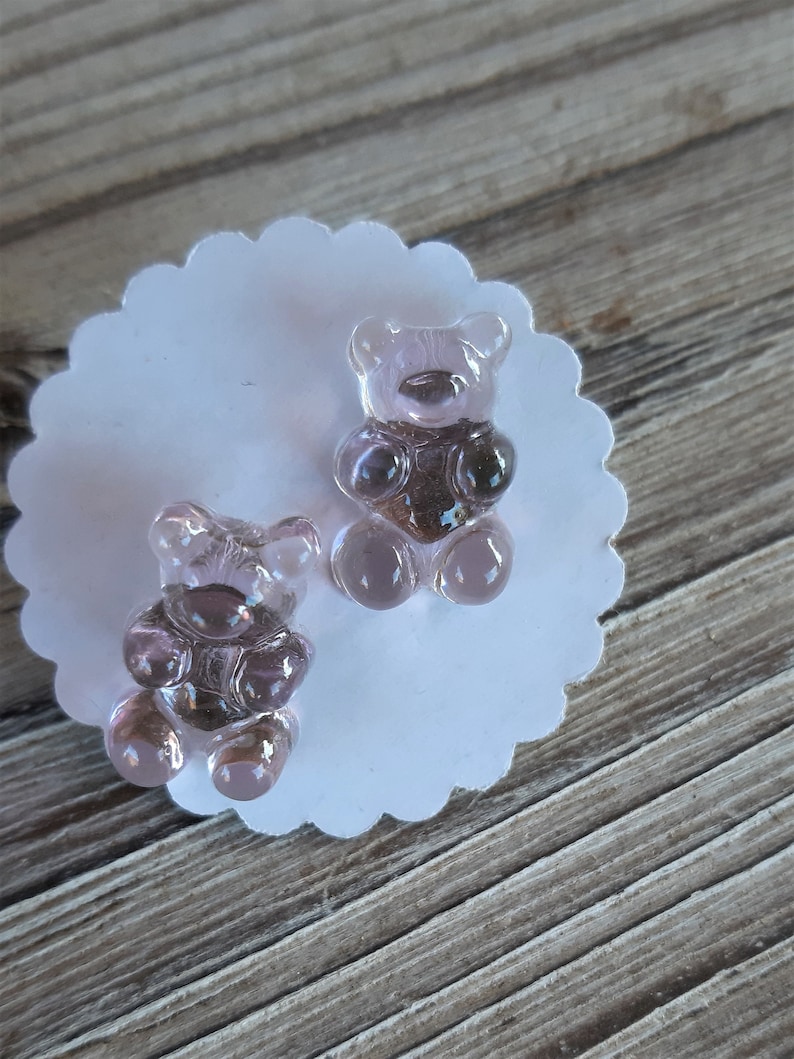 May include: Two translucent, light purple gummy bear-shaped charms sit on a white, scalloped-edge paper. The bears have a darker purple center and are positioned on a wooden surface.