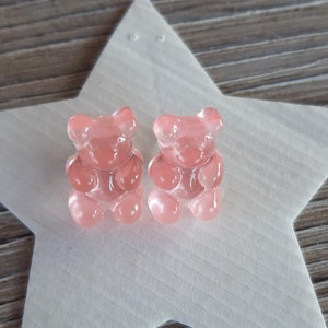 May include: Two translucent pink gummy bear candies sit on a white star-shaped surface. The gummy bears are detailed with visible features. The background is a wooden surface.