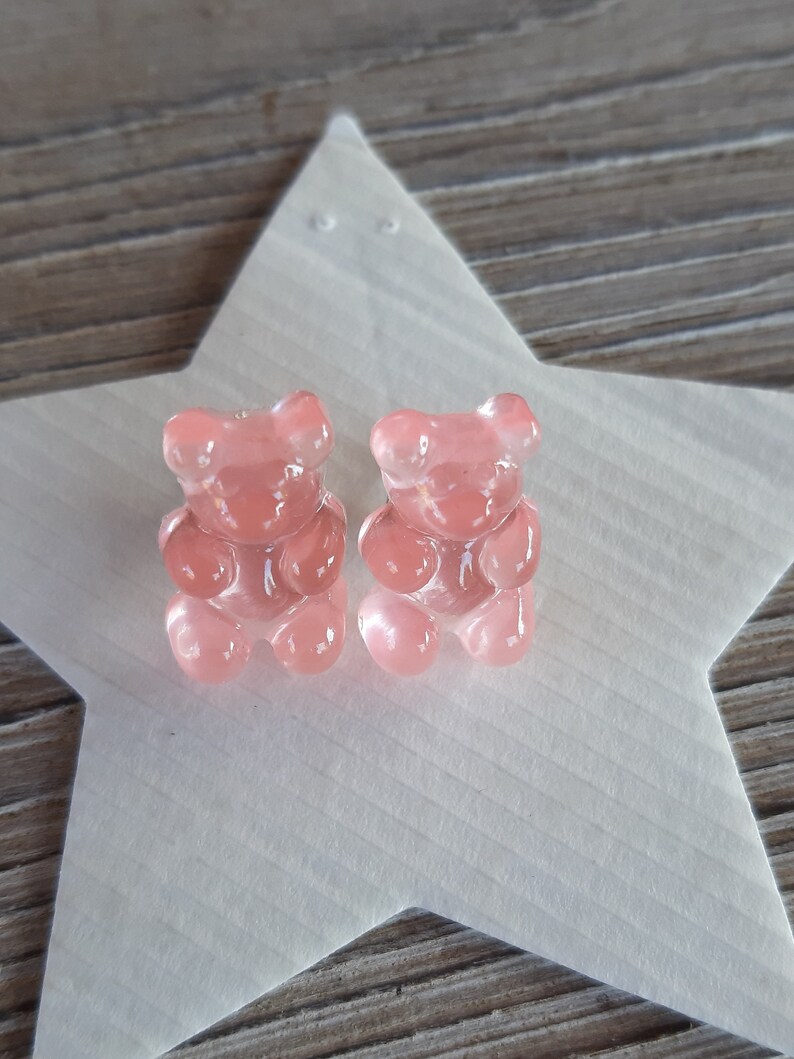 May include: Two translucent pink gummy bear candies sit on a white star-shaped surface. The gummy bears are detailed with visible features. The background is a wooden surface.