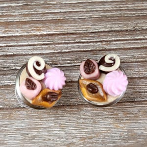May include: A pair of round stud earrings featuring miniature sculpted confections. Each earring displays a light pink swirl, a brown chocolate piece with a white spiral, and a tan pastry shape topped with a dark brown coffee bean detail.