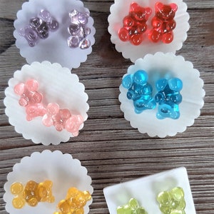 May include: A collection of gummy bear-shaped earrings in various colors, including purple, red, pink, blue, yellow, and green. Each pair of earrings is displayed on a white, scalloped paper backing, with a wooden background.