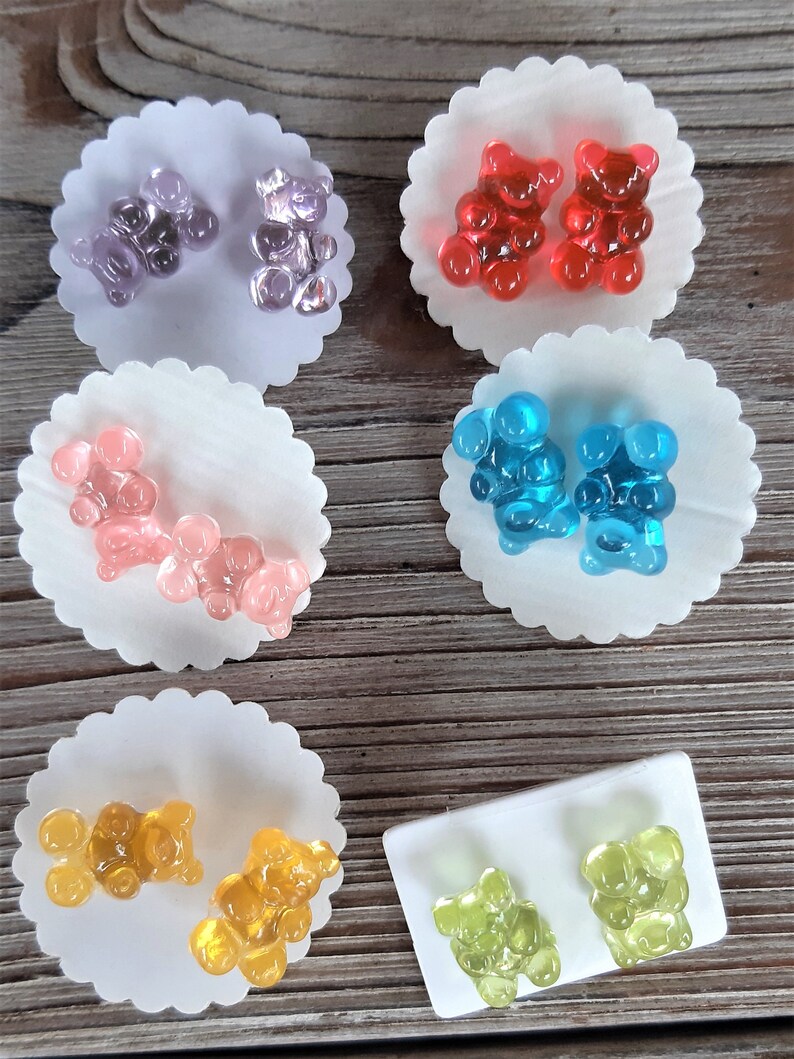 May include: A collection of gummy bear-shaped earrings in various colors, including purple, red, pink, blue, yellow, and green. Each pair of earrings is displayed on a white, scalloped paper backing, with a wooden background.