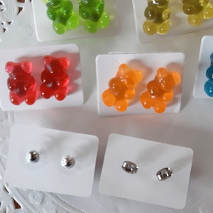 May include: A collection of colorful gummy bear earrings in green, yellow, red, orange, and blue. Also included are pairs of silver stud earrings. The earrings are displayed on white cards.