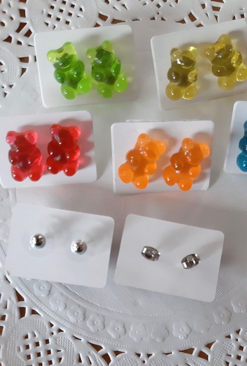 May include: A collection of colorful gummy bear earrings in green, yellow, red, orange, and blue. Also included are pairs of silver stud earrings. The earrings are displayed on white cards.