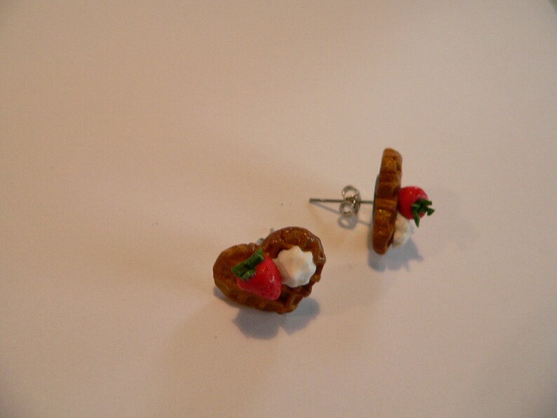 Puede incluir: Un par de pendientes de bot&oacute;n hechos a mano con forma de gofres en miniatura. Cada pendiente presenta una fresa roja y crema blanca. Los gofres son marrones con una superficie texturizada y los pendientes tienen postes plateados.