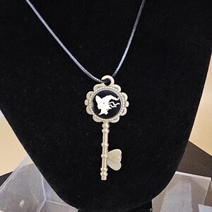 May include: A silver-colored key pendant necklace. The key has a black and white cameo design with a silhouette of a woman's face. The pendant hangs from a thin, black cord. The key is displayed on a black velvet bust.